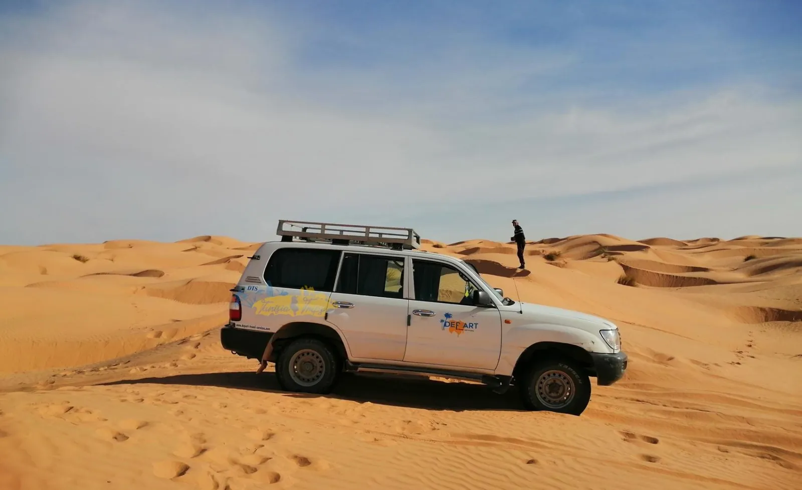 A tour 4x4 parked on the sand with a walker on the crest beyond